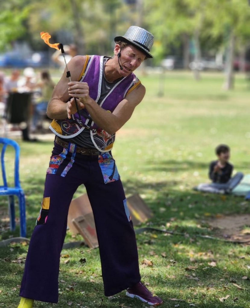 מופע ג'אגלינג של קרקס בגיל. חובש כובע צילינדר כסוף ותלבושת צבעונית, מרוכז בפעלול מרשים עם לפיד אש, במהלך מופע ג'אגלינג באוויר הפתוח בפארק.